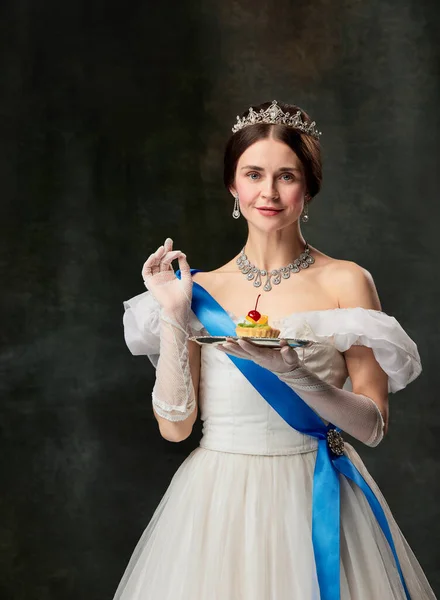 Sweet delight. Elegant woman, royal person, queen or princess in white medieval outfit tasting cake on dark vintage background. Concept of comparison of eras, modern sweets, fashion, beauty, ad