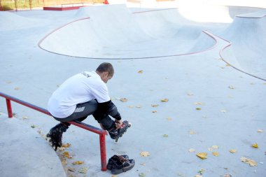 Beyaz tişörtlü ve kot pantolonlu genç adam rampada oturuyor ve paten takıyor. Hafta sonu antrenmanı. Spor anlayışı, hobiler, aktif sağlıklı yaşam tarzı..