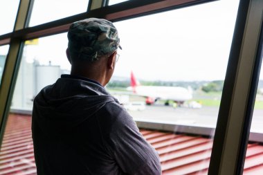 Havaalanındaki büyük pencerelerden uçağa bakan genç adam uçağa binmeyi bekliyor..