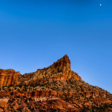 Beautiful rock formations in Arizona, US. Prominent orange butte, with contrasting green plants and blue sky. The moon is visible on top