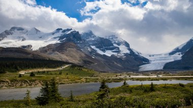 Stunning sun rays shining on the peaks and glaciers at Columbia Ice field, Alberta, Canada. Beautiful light coming from the dramatic sky. Green trees, grass and a river in the foreground