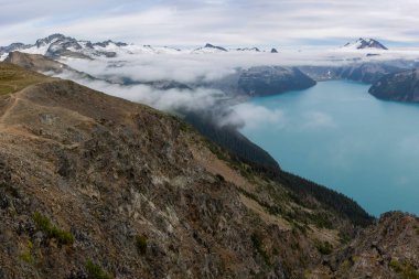 Kanada, British Columbia 'daki Panorama Ridge yürüyüşünden Garibaldi Gölü' nün güzel manzarası. Dağlar ve buzullar arka plandaki bulutların arasından gözetliyor. Turkuaz göl bulutların altında.