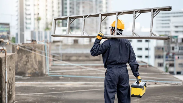 Asian maintenance worker man with protective suit and safety helmet carrying aluminium step ladder and tool box at construction site. Civil engineering, Architecture builder and building service