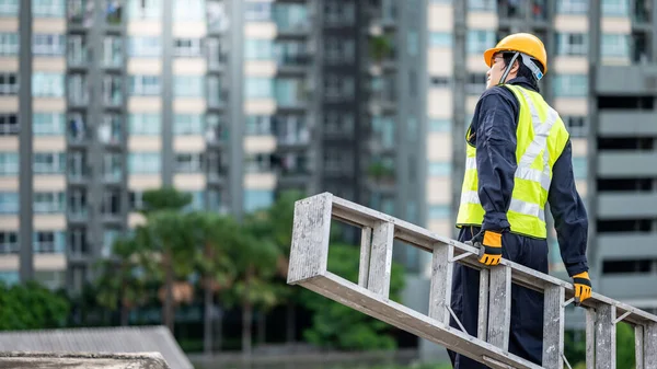 Asian maintenance worker man with safety helmet and green vest carrying aluminium step ladder at construction site. Civil engineering, Architecture builder and building service concepts