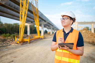 İnşaat alanında proje planlama ve mimari çizimleri için dijital tablet kullanan koruyucu kasklı ve yansıtıcı yelekli akıllı Asyalı işçi ya da erkek inşaat mühendisi..