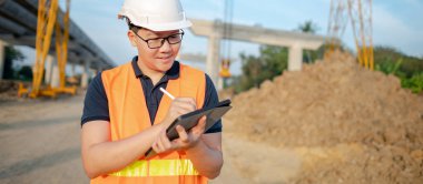 İnşaat alanında proje planlama ve mimari çizimleri için dijital tablet kullanan koruyucu kasklı ve yansıtıcı yelekli akıllı Asyalı işçi ya da erkek inşaat mühendisi..