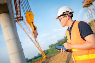 İnşaat alanında proje planlama ve mimari çizimleri için dijital tablet kullanan koruyucu kasklı ve yansıtıcı yelekli akıllı Asyalı işçi ya da erkek inşaat mühendisi..