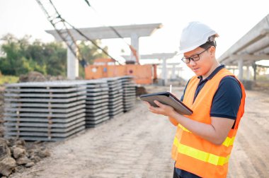 İnşaat alanındaki projeleri planlamak ve kontrol etmek için dijital tablet kullanan koruyucu kasklı ve yansıtıcı yelekli akıllı Asyalı işçi ya da erkek inşaat mühendisi..