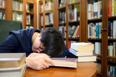 Asyalı bir üniversite öğrencisi, eğitim ve araştırma konusu için üniversite kütüphanesinde kitaplığın yanında kitap okurken uyuyor. Eğitim fırsatı için burs.
