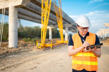 İnşaat alanında proje planlama ve mimari çizimleri için dijital tablet kullanan koruyucu kasklı ve yansıtıcı yelekli akıllı Asyalı işçi ya da erkek inşaat mühendisi..