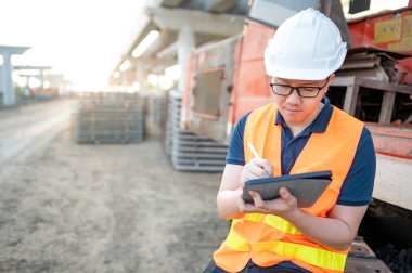 İnşaat alanındaki proje planını kontrol etmek için dijital tablet kullanan kazıcı makinesinin üzerinde koruyucu kaskı ve yansıtıcı yeleği olan akıllı Asyalı işçi veya erkek inşaat mühendisi..