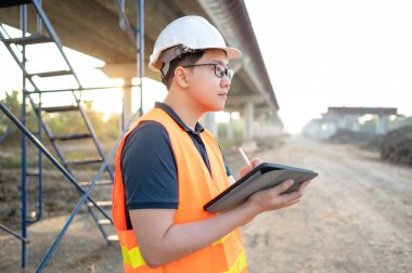 İnşaat alanında proje planlama ve mimari çizimleri için dijital tablet kullanan koruyucu kasklı ve yansıtıcı yelekli akıllı Asyalı işçi ya da erkek inşaat mühendisi..