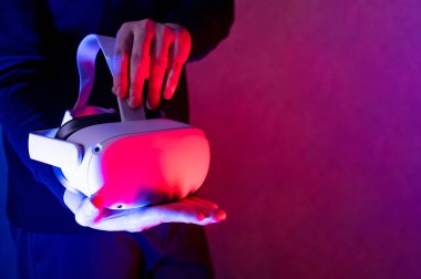 Erkek oyuncu elleri, neon ışıklı odada sanal gerçeklik gözlüklerini tutuyor. Eğlence deneyimi için VR kulaklık teknolojisi. Metaverse görselleştirme simülasyonu, 3D, AR, VR, Innovation.