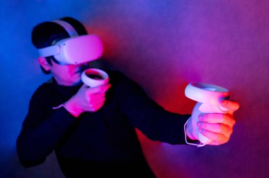 Sanal gerçeklik gözlüğü takan Asyalı oyuncu neon ışıklı stüdyoda video oyunları oynayan kontrolörleri tutuyor. Eğlence deneyimi için VR kulaklık teknolojisi. Şekillendirme canlandırma simülasyonu kavramı