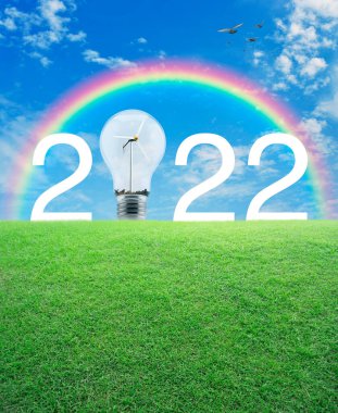 Gökkuşağı üzerinde yeşil çimen tarlasında rüzgar türbini bulunan 2022 beyaz metin ve ampul, beyaz bulutlu kuşlar ve mavi gökyüzü, mutlu yeni yıl 2022 ekolojik kapak kavramı