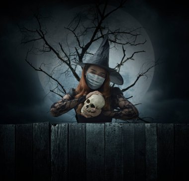 Ölü ağacın, dolunayın ve ürkütücü bulutlu gökyüzünün, Cadılar Bayramı 'nın ve Coronavirüs' ün ya da covid-19 konseptinin üzerinde duran bir kafatası tutan tıbbi maske takan cadılar bayramı cadısı.