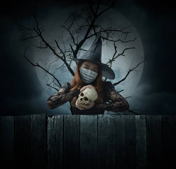 Ölü ağacın, dolunayın ve ürkütücü bulutlu gökyüzünün, Cadılar Bayramı 'nın ve Coronavirüs' ün ya da covid-19 konseptinin üzerinde duran bir kafatası tutan tıbbi maske takan cadılar bayramı cadısı.