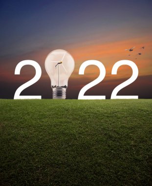 2022 beyaz yazı ve ampul içinde rüzgar türbini olan yeşil çimlerin üzerinde günbatımı gökyüzünde kuşlarla birlikte. Mutlu yıllar 2022 ekolojik kapak kavramı.