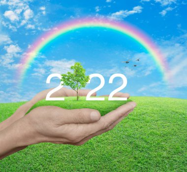 2022 beyaz metin yeşil çimen tarlasında büyüyen ağaç insan ellerinde mavi gökyüzü, beyaz bulutlar ve gökkuşağı, mutlu yeni yıl 2022 ekolojik kapak konsepti