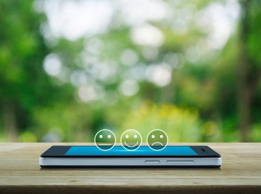 Parktaki bulanık yeşil ağacın üzerinde, ahşap masadaki modern akıllı cep telefonu ekranında gülen surat ikonu, iş dünyası müşteri hizmetleri değerlendirmesi ve çevrimiçi değerlendirme konsepti