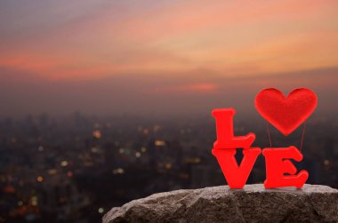 Kırmızı aşk alfabesi ve kumaştan kalp balonu şehir manzarasının üzerinde, sıcak gün batımında, Sevgililer Günü konseptinde.