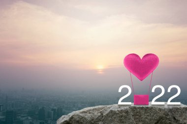 Pembe kumaşlı kalp, 2022 beyaz yazıyla hava balonuna bayılıyor. Gün batımında şehir manzarasının havadan görünüşü, klasik tarz, mutlu yıllar ve 2022 Sevgililer Günü konsepti.