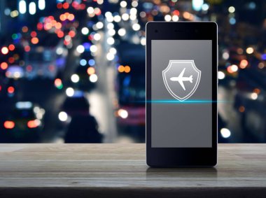 Kalkan düz ikona sahip uçak modern akıllı cep telefonu ekranında ahşap masa üzerinde bulanık renkli gece lambası trafik sıkışıklığı yolu üzerinde şehirde arabalar, iş seyahati sigortası ve güvenlik online konsepti