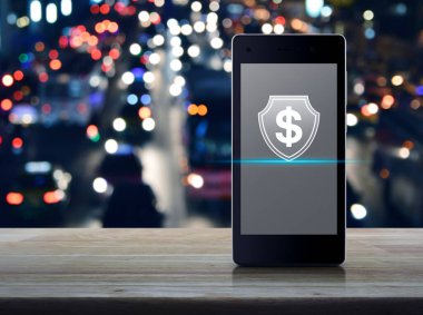 Kalkan düz simgeli dolar, modern akıllı cep telefonu ekranında ahşap masa üzerinde bulanık renkli gece lambası trafik sıkışıklığı yolu üzerinde, şehirde arabalar, iş parası sigortası ve çevrimiçi koruma kavramı ile.