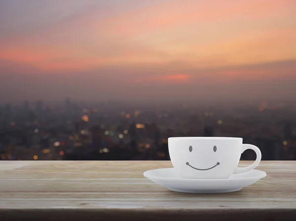 White coffee cup with smile face shape on wooden table over blur of cityscape on warm light sundown