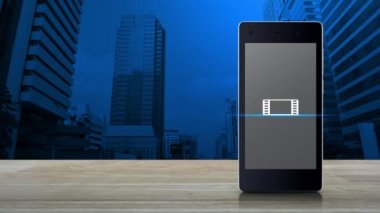 Ofis kulesi ve gökdelenin üzerindeki ahşap masada modern akıllı cep telefonu ekranında film simgesi ile oynat, iş sineması çevrimiçi konsepti