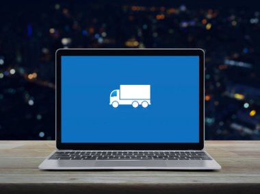Delivery truck flat icon on modern laptop computer screen on wooden table over blur colorful night light office city tower and skyscraper, Business transportation online service concept