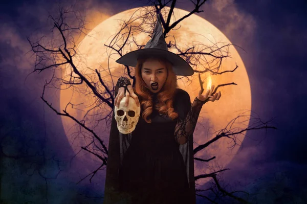 Halloween witch holding a skull standing over dead tree, full moon and spooky cloudy sky, Halloween mystery concept