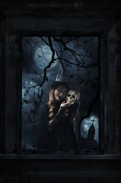 Halloween witch holding a skull standing in old damaged wood window with wall over cross, church, birds, dead tree and spooky cloudy sky, Halloween mystery concept