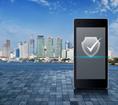 Security shield with check mark icon on modern smart mobile phone screen on stone tile floor over city tower, river and blue sky, Technology internet cyber security online concept
