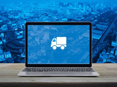 Delivery truck flat icon on modern laptop computer screen on wooden table over city tower, street, expressway and skyscraper, Business transportation online service concept