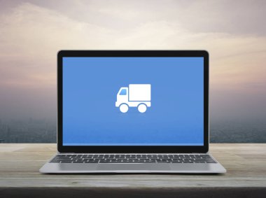 Delivery truck flat icon on modern laptop computer screen on wooden table over office city tower and skyscraper at sunset sky, vintage style, Business transportation online service concept