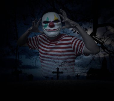 Scary evil clown standing over grass, dead tree, cross, birds with church over spooky cloudy sky, Halloween mystery concept