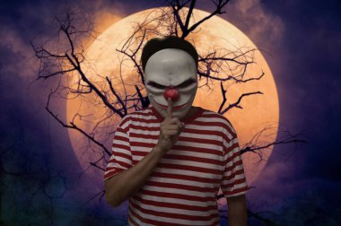 Scary evil clown showing silence sign with finger over lips standing over dead tree, full moon and spooky cloudy sky, Halloween mystery concept
