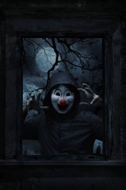 Scary evil clown wear jacket standing in old damaged wood window with wall over cross, church, birds, dead tree, full moon and spooky cloudy sky, Halloween mystery concept
