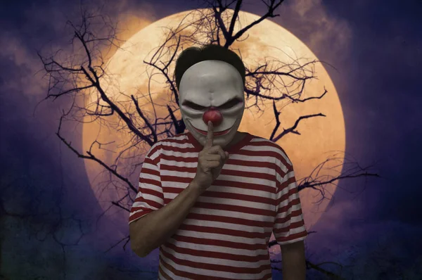 Scary evil clown showing silence sign with finger over lips standing over dead tree, full moon and spooky cloudy sky, Halloween mystery concept