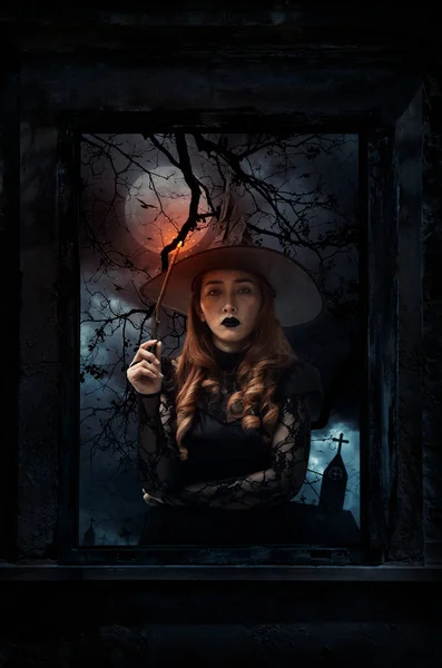 Halloween witch holding magic wand standing in old damaged wood window with wall over cross, church, birds, dead tree and spooky cloudy sky, Halloween mystery concept