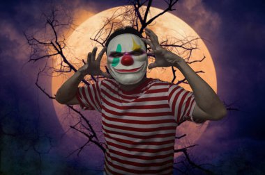 Ölü ağacın üzerinde duran korkunç kötü palyaço, dolunay ve ürkütücü bulutlu gökyüzü, Cadılar Bayramı gizemi kavramı