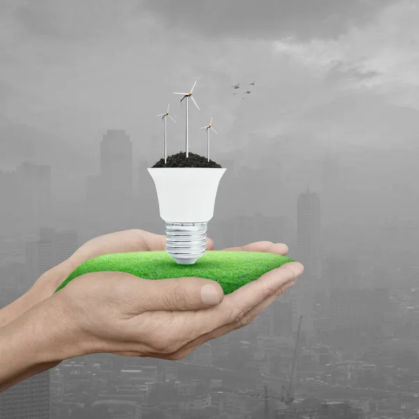 Wind turbines on soil with light bulb on green grass in hands over pollution city tower and skyscraper with birds, Green ecology and saving energy concept