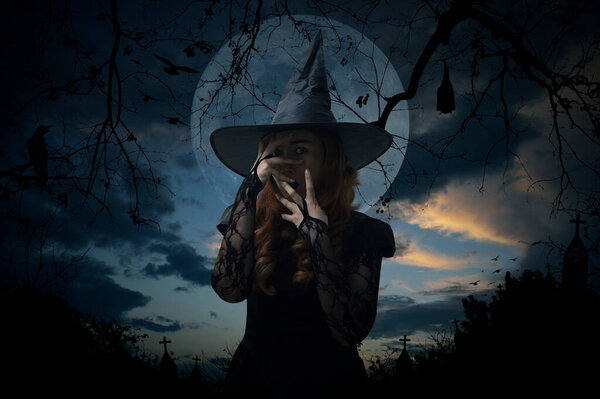 Scary halloween witch standing over cross, church, crow, bat, birds, dead tree, full moon and sunset sky, Halloween mystery concept