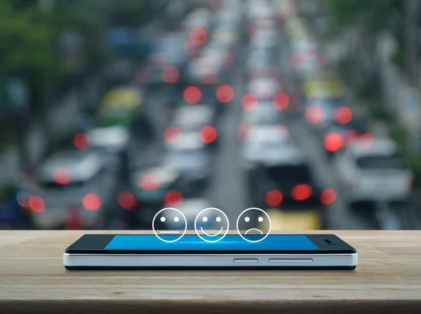 Excellent smiley face rating icon on modern smart mobile phone screen on wooden table over blur of rush hour with cars and road in city, Business customer service evaluation and feedback rating online concept