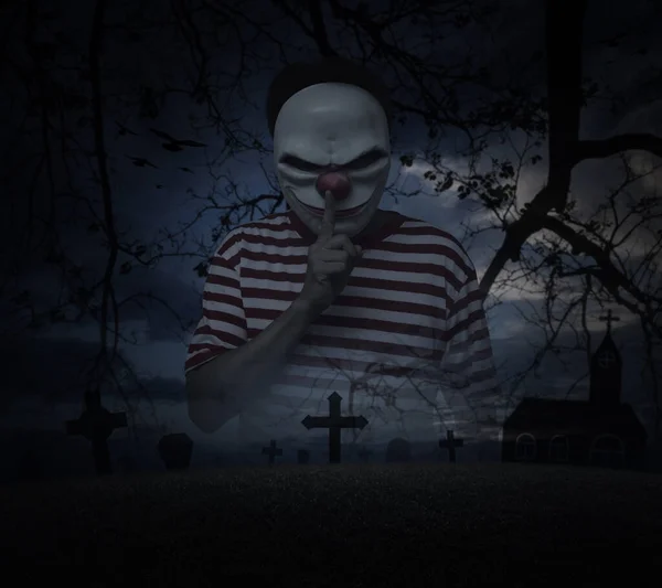 Scary evil clown showing silence sign with finger over lips standing over grass, dead tree, cross, church and spooky cloudy sky, Halloween mystery concept