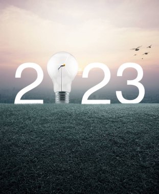2023 beyaz yazı ve ampul, içinde rüzgar türbini olan yeşil çimen tarlasında günbatımında şehir manzarası, klasik tarz, mutlu yeni yıl ekolojik kapak konsepti.