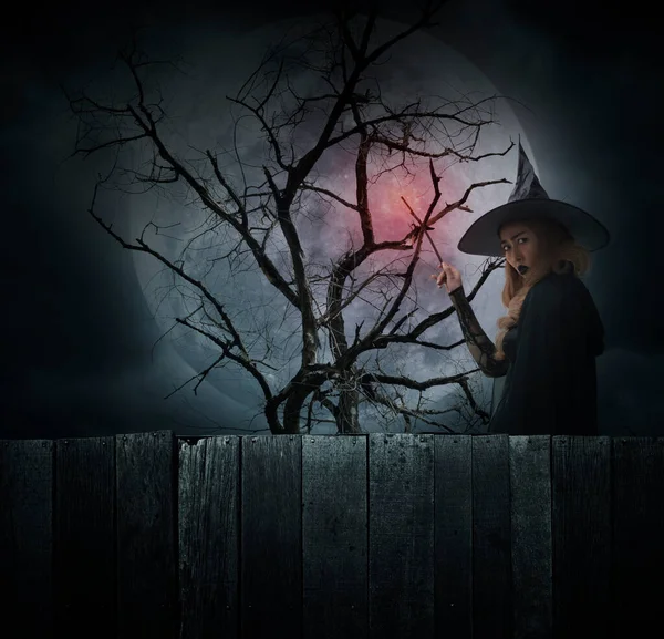 Halloween witch holding magic wand standing over dead tree, full moon and cloudy spooky sky, Halloween mystery concept