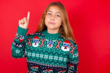 Noel süveteri giyen güzel bir kız kendini beğenmiş ve mutlu bir şekilde gülümserken parmaklarıyla bir numarayı gösteriyor..