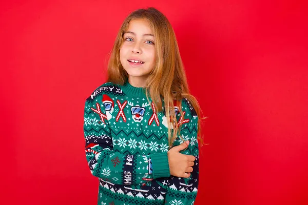 赤い背景のスタジオで笑うクリスマスセーターを着ている美しい女の子 — ストック写真